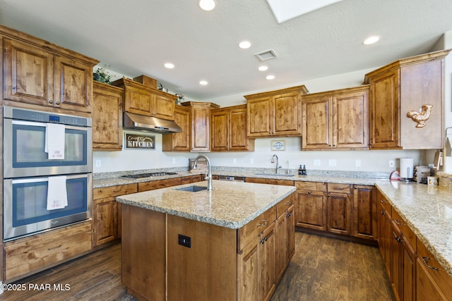 kitchen with double oven, dark wood-style floors, under cabinet range hood, brown cabinets, and a sink
