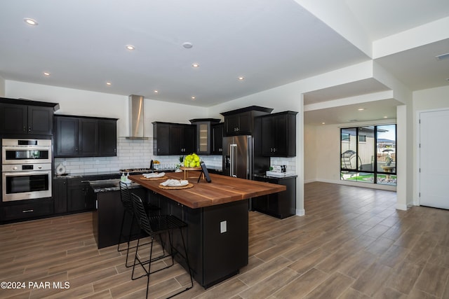 kitchen with a center island, stainless steel appliances, wall chimney range hood, wooden counters, and hardwood / wood-style floors