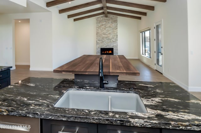 kitchen with dark stone counters, sink, lofted ceiling with beams, hardwood / wood-style floors, and a stone fireplace