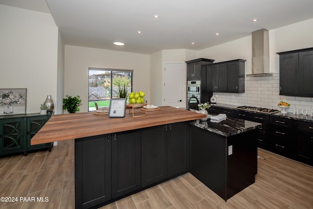 kitchen with wooden counters, appliances with stainless steel finishes, light wood-type flooring, wall chimney exhaust hood, and a center island with sink