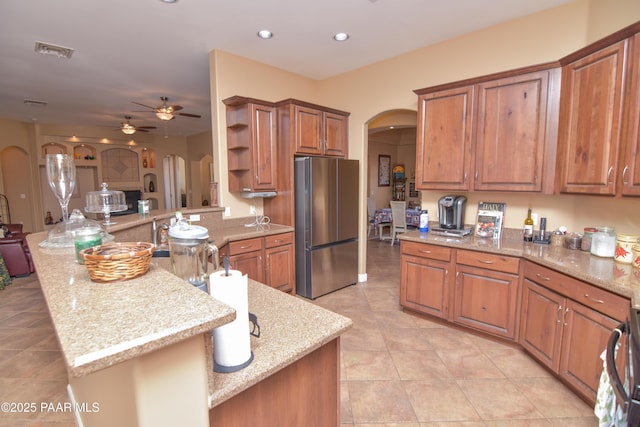 kitchen with arched walkways, freestanding refrigerator, a peninsula, ceiling fan, and brown cabinetry