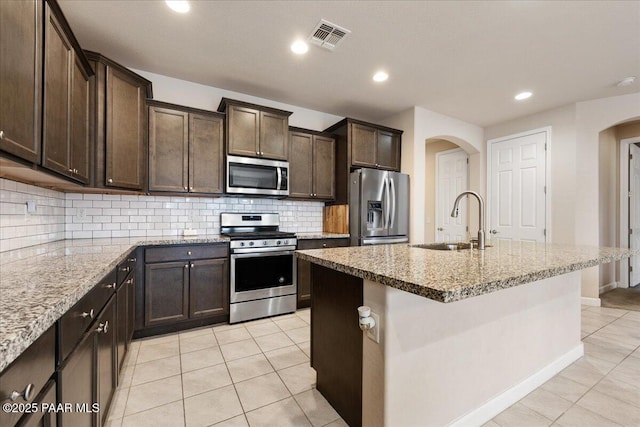 kitchen with sink, light tile patterned floors, stainless steel appliances, light stone countertops, and an island with sink