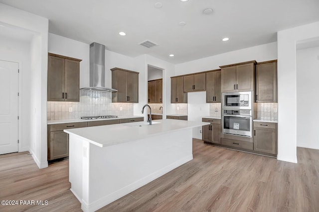 kitchen with wall chimney range hood, sink, light hardwood / wood-style flooring, appliances with stainless steel finishes, and a center island with sink