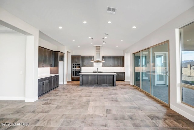 kitchen with wall chimney range hood, sink, backsplash, an island with sink, and vaulted ceiling