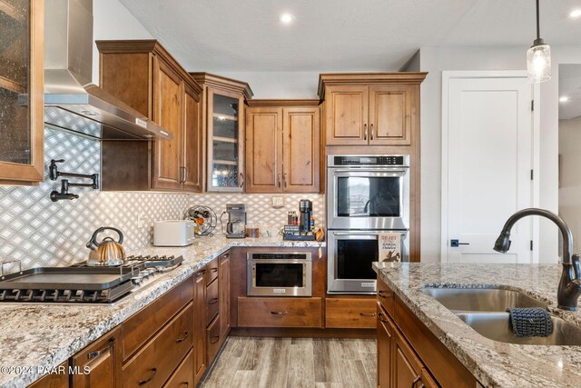 kitchen with appliances with stainless steel finishes, light wood-type flooring, sink, wall chimney range hood, and hanging light fixtures