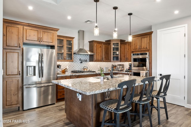 kitchen with stainless steel appliances, wall chimney range hood, light stone counters, wood-type flooring, and a center island with sink