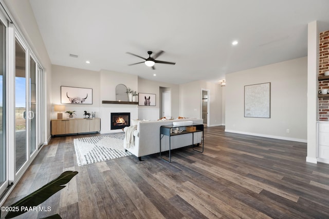 living area featuring visible vents, dark wood-type flooring, a glass covered fireplace, and ceiling fan