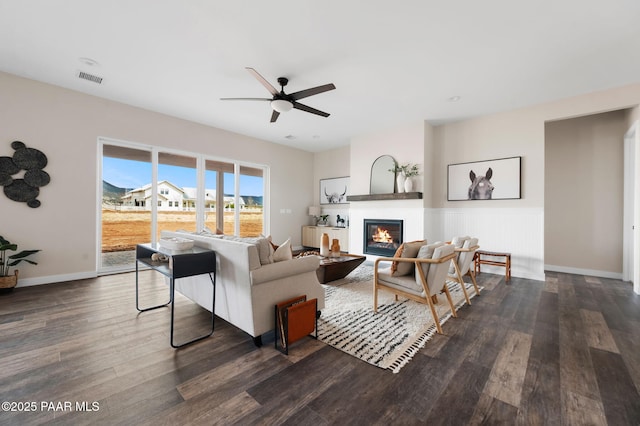 living area with dark wood-type flooring, a wainscoted wall, visible vents, a glass covered fireplace, and ceiling fan