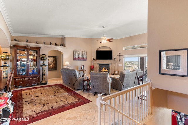 living room featuring ceiling fan, ornamental molding, tile patterned floors, arched walkways, and a lit fireplace