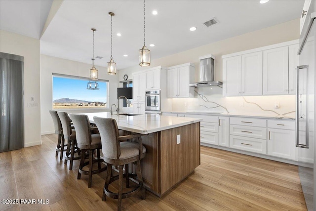 kitchen featuring a center island with sink, tasteful backsplash, white cabinets, a sink, and wall chimney range hood