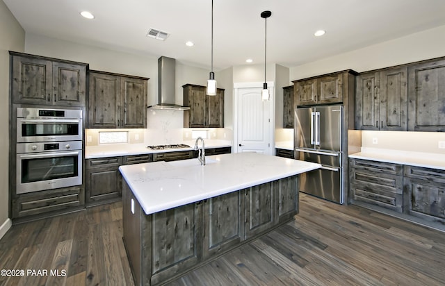 kitchen with wall chimney range hood, dark wood-type flooring, appliances with stainless steel finishes, a kitchen island with sink, and hanging light fixtures