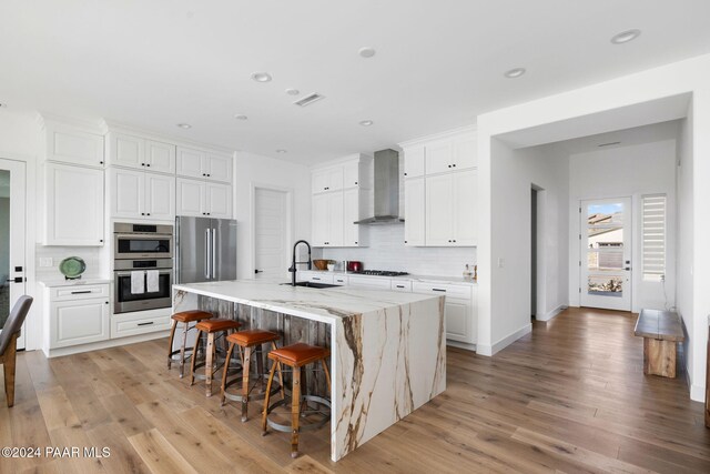 kitchen featuring light wood-type flooring, wall chimney exhaust hood, sink, a center island with sink, and white cabinets