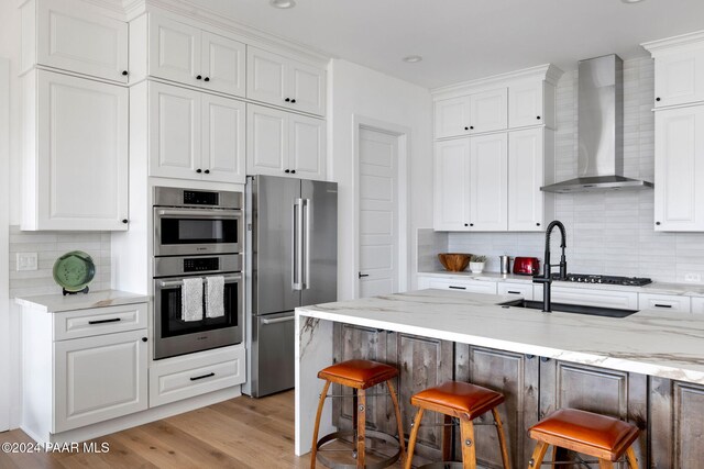 kitchen featuring wall chimney exhaust hood, stainless steel appliances, light hardwood / wood-style flooring, white cabinetry, and a breakfast bar area