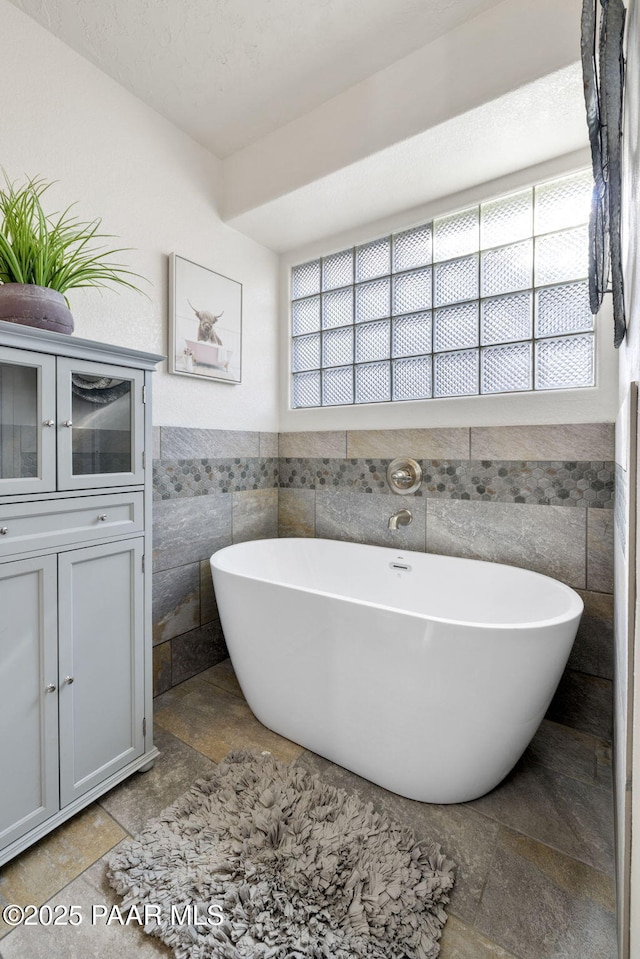 bathroom with tile walls, a soaking tub, wainscoting, and stone tile flooring