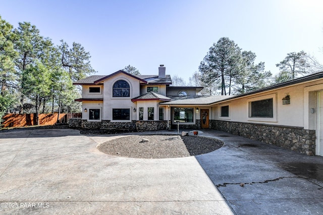 rear view of house featuring fence, concrete driveway, stone siding, stucco siding, and a chimney