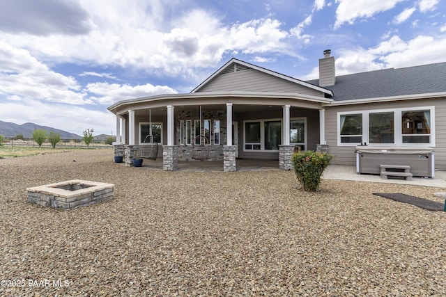 back of property featuring a hot tub, a patio area, a mountain view, a chimney, and an outdoor fire pit