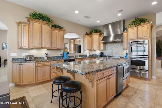 kitchen with a kitchen island with sink, wall chimney exhaust hood, dark stone countertops, appliances with stainless steel finishes, and a kitchen bar