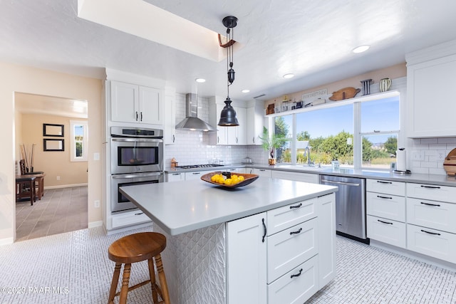 kitchen with appliances with stainless steel finishes, wall chimney range hood, white cabinets, a kitchen island, and hanging light fixtures