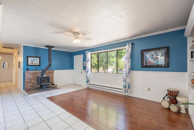 unfurnished living room featuring tile patterned flooring, ceiling fan, a wainscoted wall, a wood stove, and a baseboard radiator