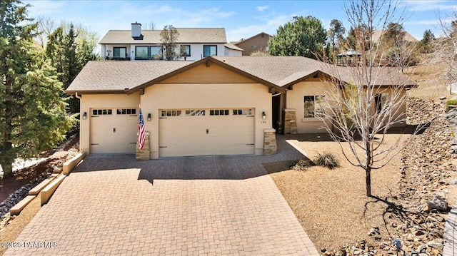 ranch-style house with a shingled roof, an attached garage, stone siding, stucco siding, and decorative driveway