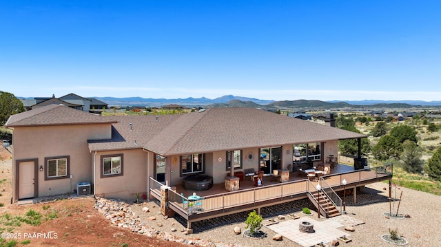 rear view of property with a deck with mountain view, a patio, a fire pit, a shingled roof, and stucco siding