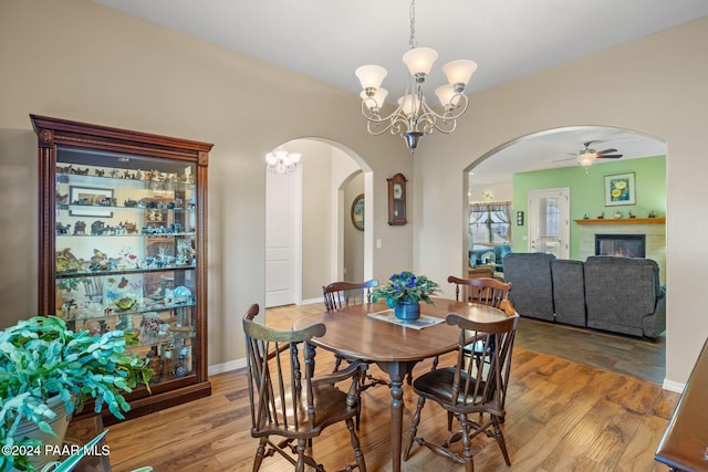 dining room with ceiling fan with notable chandelier, wood finished floors, arched walkways, a lit fireplace, and baseboards
