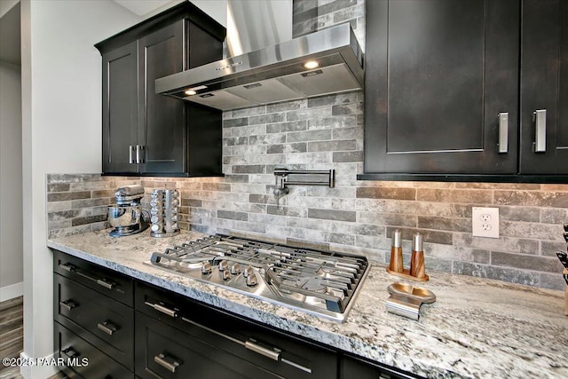 kitchen with light stone counters, wall chimney exhaust hood, baseboards, decorative backsplash, and stainless steel gas stovetop