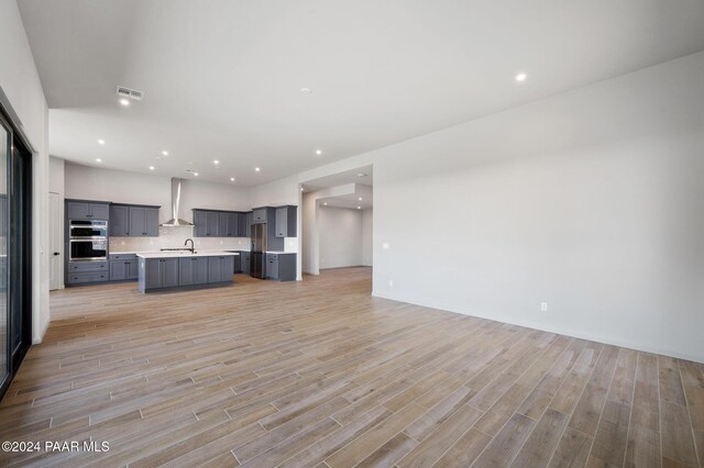 kitchen with backsplash, wall chimney exhaust hood, double oven, a center island with sink, and light hardwood / wood-style flooring