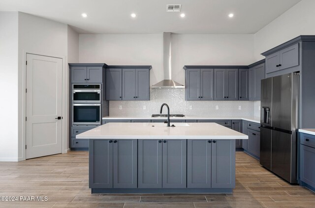 kitchen featuring wall chimney range hood, stainless steel appliances, a kitchen island with sink, and light hardwood / wood-style flooring