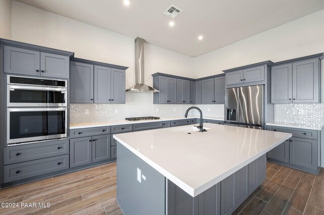 kitchen with dark hardwood / wood-style flooring, wall chimney exhaust hood, a center island with sink, and appliances with stainless steel finishes