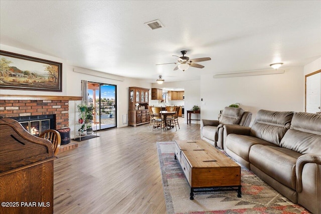 living area featuring light wood finished floors, visible vents, ceiling fan, a textured ceiling, and a brick fireplace