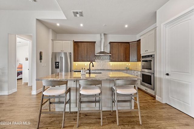 kitchen featuring wall chimney exhaust hood, a breakfast bar, stainless steel appliances, a kitchen island with sink, and light hardwood / wood-style floors