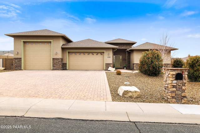 prairie-style home with stucco siding, roof with shingles, an attached garage, stone siding, and decorative driveway