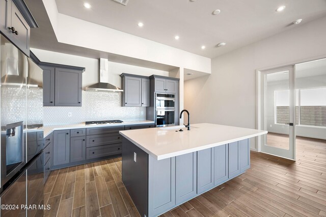 kitchen featuring sink, appliances with stainless steel finishes, backsplash, an island with sink, and wall chimney exhaust hood