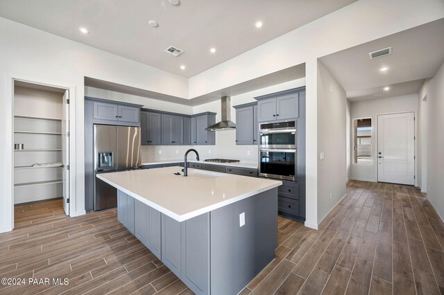 kitchen with sink, an island with sink, stainless steel appliances, decorative backsplash, and wall chimney range hood