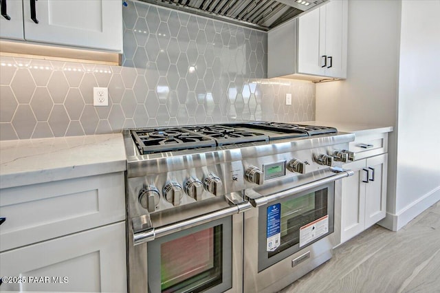 kitchen featuring light stone counters, tasteful backsplash, white cabinets, range with two ovens, and ventilation hood
