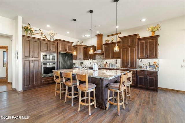 kitchen featuring a kitchen island with sink, dark wood finished floors, stainless steel appliances, wall chimney exhaust hood, and decorative backsplash