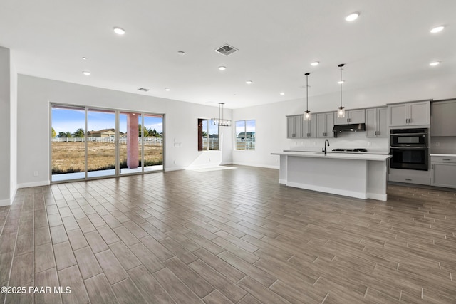 kitchen with double wall oven, hanging light fixtures, a center island with sink, and gray cabinetry