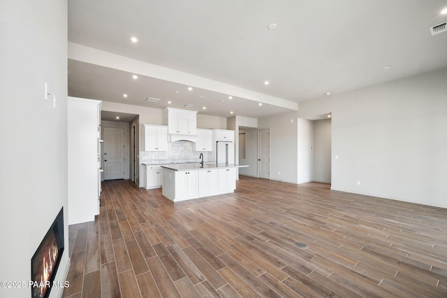 kitchen featuring open floor plan, wood finished floors, a kitchen island with sink, light countertops, and backsplash