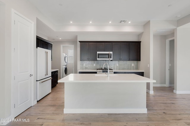 kitchen with stacked washer / drying machine, high end appliances, light wood-type flooring, and an island with sink