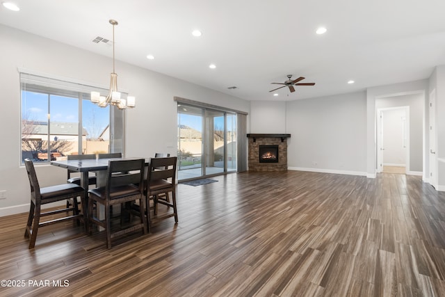 dining area featuring dark wood-type flooring, ceiling fan with notable chandelier, a healthy amount of sunlight, and a stone fireplace