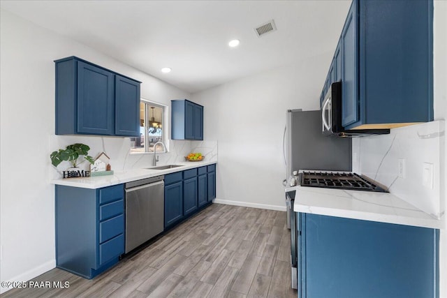 kitchen with light stone countertops, stainless steel appliances, sink, blue cabinetry, and light hardwood / wood-style floors