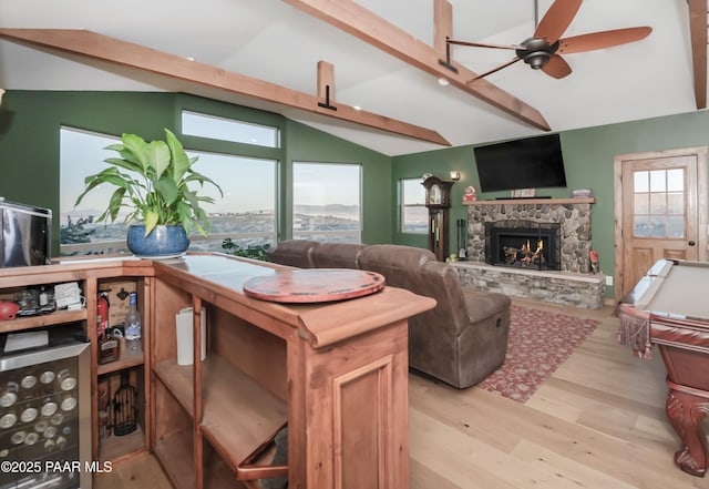 living room with ceiling fan, vaulted ceiling with beams, a wealth of natural light, a stone fireplace, and light wood-type flooring