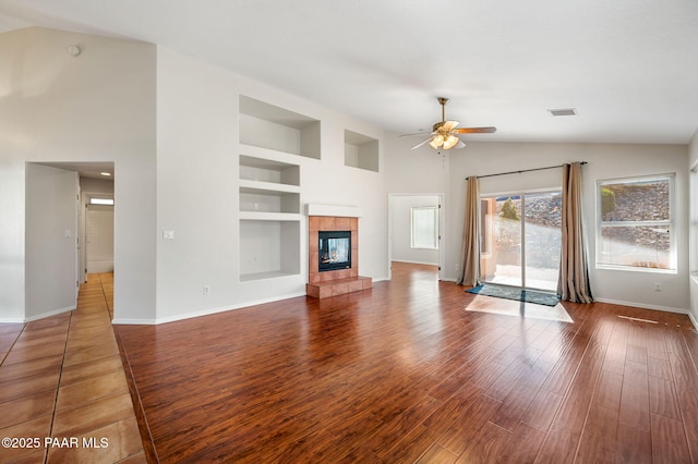 unfurnished living room featuring built in features, high vaulted ceiling, hardwood / wood-style flooring, a tiled fireplace, and ceiling fan