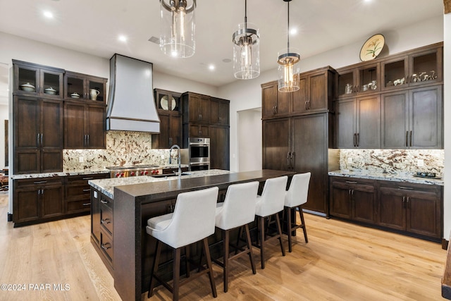 kitchen with backsplash, premium range hood, sink, hanging light fixtures, and an island with sink