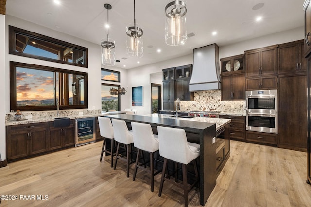 kitchen featuring wine cooler, decorative backsplash, pendant lighting, a kitchen island with sink, and custom range hood