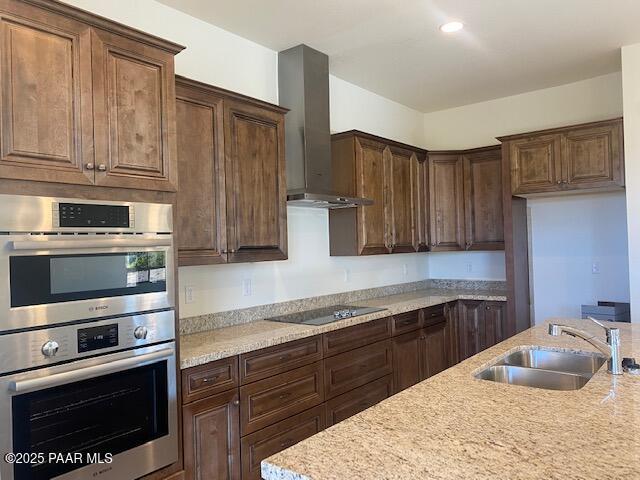 kitchen featuring sink, wall chimney exhaust hood, stainless steel double oven, light stone counters, and black electric cooktop