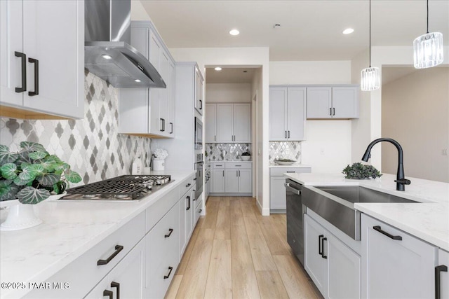 kitchen featuring wall chimney exhaust hood, light stone counters, decorative light fixtures, stainless steel appliances, and a sink