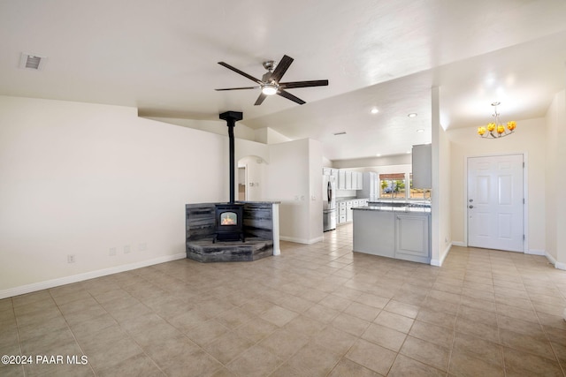 unfurnished living room with baseboards, visible vents, a wood stove, vaulted ceiling, and ceiling fan with notable chandelier