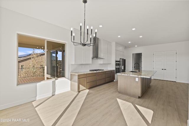 kitchen featuring wall chimney exhaust hood, a large island with sink, decorative light fixtures, light hardwood / wood-style floors, and white cabinetry